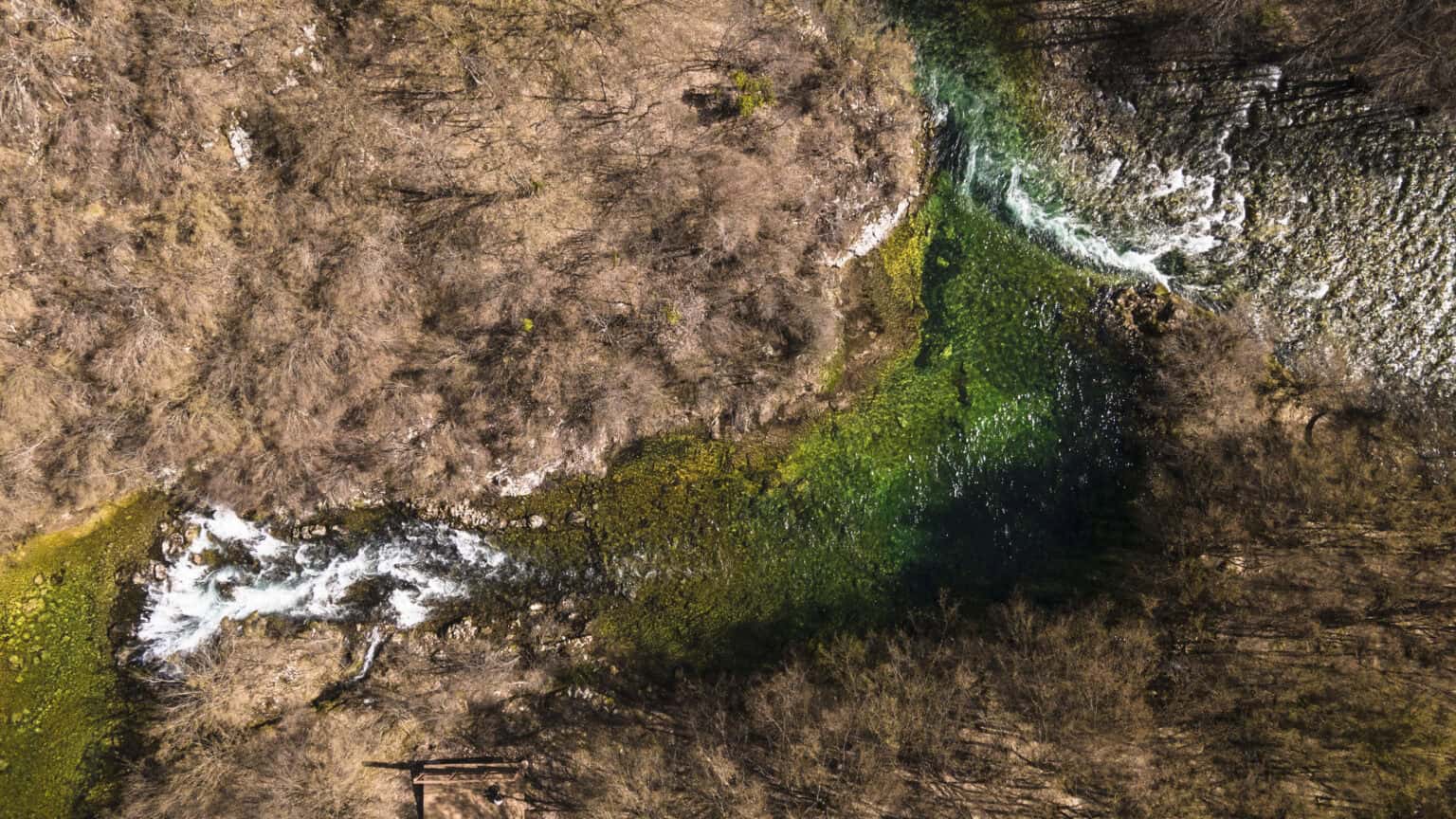 Pogled iz zraka na rijeku Sanu i njene bistre zelene tokove okružene suhom vegetacijom, simbol netaknute prirode Bosne i Hercegovine.