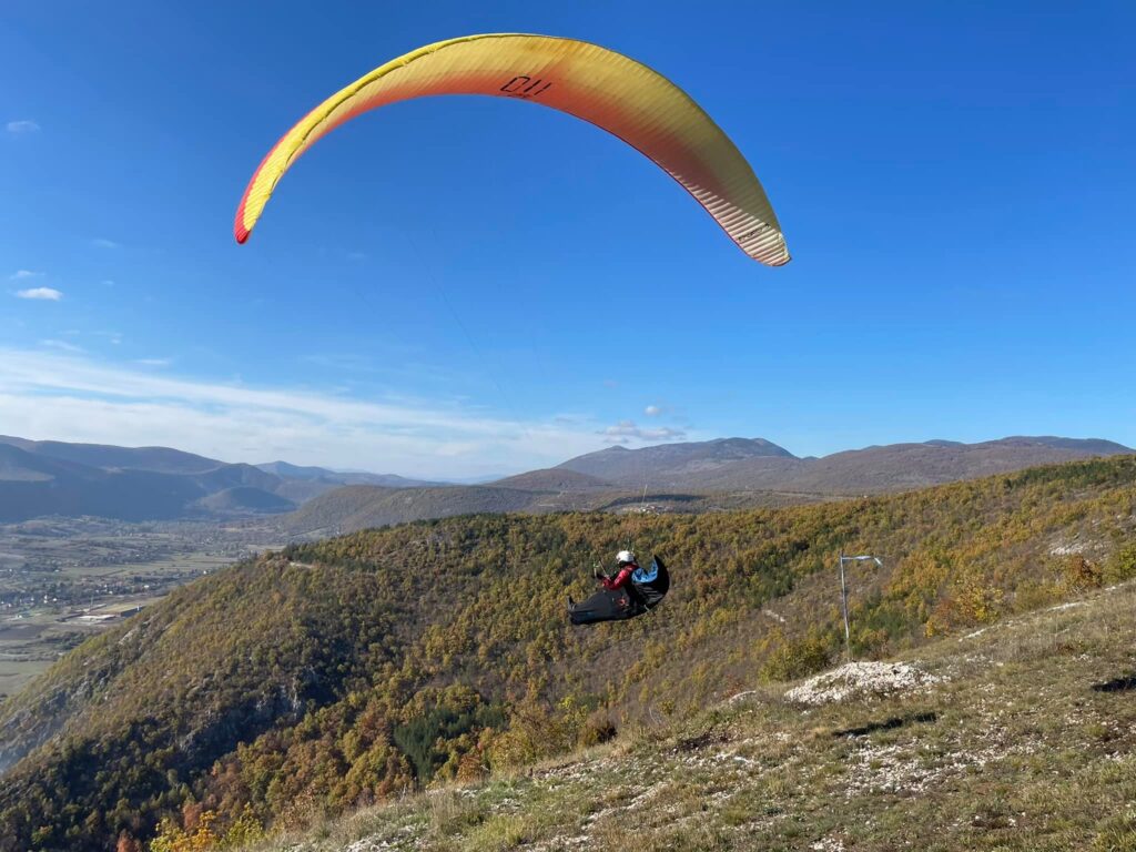 Paraglajder u letu iznad brdovitog pejzaža Livanjskog polja, okružen jesenjim bojama i plavim nebom.