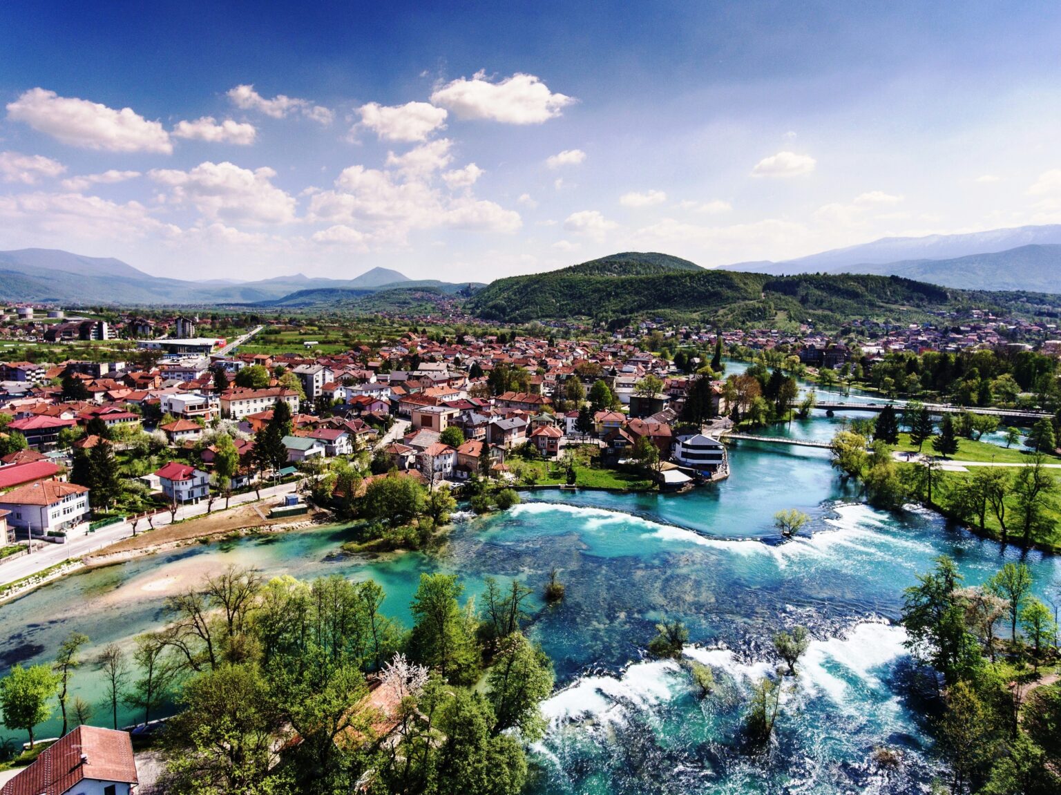 Panoramski pogled na Bihać i rijeku Unu s kristalno čistom vodom, slapovima i zelenilom u pozadini.