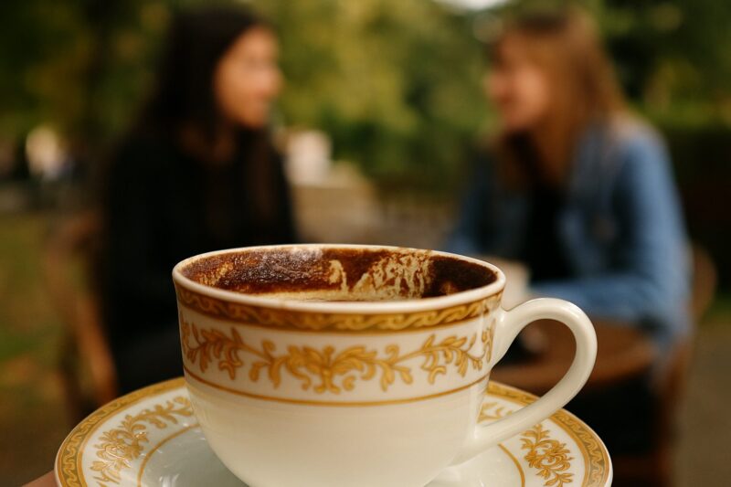 Tradicionalna šoljica kafe sa zlatnim ornamentima, u pozadini dvije osobe u razgovoru na otvorenom, simbolizirajući ritual ispijanja kafe u Krajini.